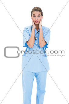 Astonished brown haired nurse in blue scrubs posing with head in hands