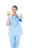 Cheering brown haired nurse in blue scrubs holding a green apple