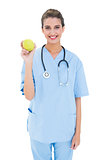 Pleased brown haired nurse in blue scrubs holding a green apple