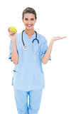 Cheerful brown haired nurse in blue scrubs holding a green apple