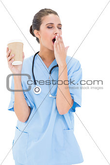 Tired brown haired nurse in blue scrubs yawning and holding a cup of coffee