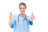 Amused brown haired nurse in blue scrubs giving a thumb up while holding a glass of water