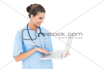 Happy brown haired nurse in blue scrubs using a laptop