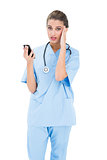 Confused brown haired nurse in blue scrubs using a mobile phone