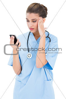 Puzzled brown haired nurse in blue scrubs using a mobile phone