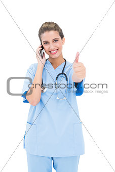 Cheerful brown haired nurse in blue scrubs making a phone call and raising her thumb