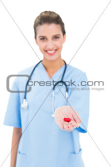 Amused brown haired nurse in blue scrubs offering a pill