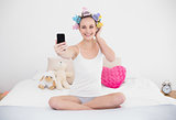 Smiling natural brown haired woman in hair curlers taking a picture of herself with mobile phone