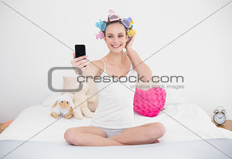 Smiling natural brown haired woman in hair curlers taking a picture of herself with mobile phone