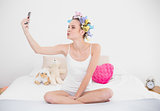 Playful natural brown haired woman in hair curlers taking a picture of herself with mobile phone