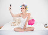 Amused natural brown haired woman in hair curlers taking a picture of herself with mobile phone