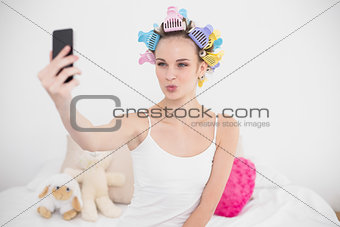Funny natural brown haired woman in hair curlers taking a picture of herself with mobile phone