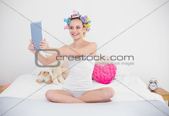 Cheerful natural brown haired woman in hair curlers taking picture of herself with a tablet