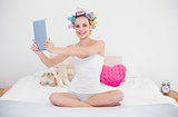 Joyful natural brown haired woman in hair curlers taking picture of herself with a tablet