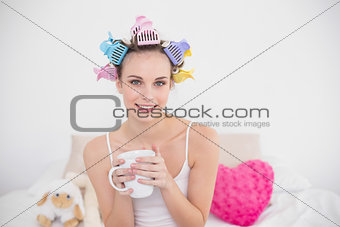 Pleased natural brown haired woman in hair curlers holding a cup of coffee