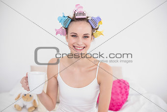 Joyful natural brown haired woman in hair curlers holding a cup of coffee
