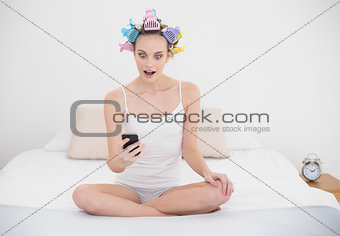 Astonished natural brown haired woman in hair curlers looking at her mobile phone