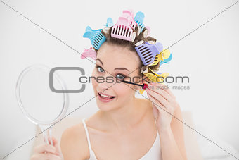 Cheerful natural brown haired woman in hair curlers applying mascara