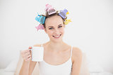 Amused natural brown haired woman in hair curlers holding a mug of coffee