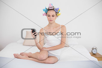 Frowning natural brown haired woman in hair curlers looking at her mobile phone