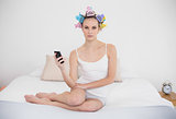Frowning natural brown haired woman in hair curlers holding a mobile phone