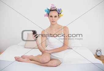 Frowning natural brown haired woman in hair curlers holding a mobile phone