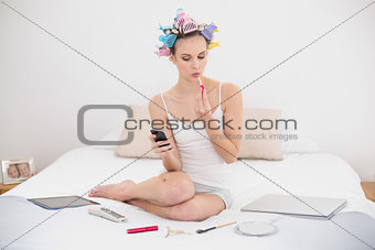 Concentrated natural brown haired woman in hair curlers applying gloss while using smartphone