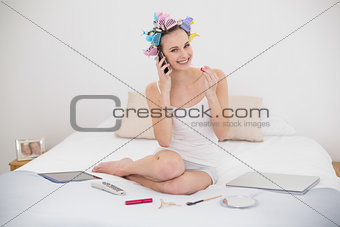 Pleased natural brown haired woman in hair curlers applying gloss while being on the phone