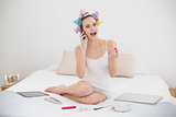 Surprised natural brown haired woman in hair curlers applying gloss while being on the phone