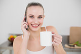 Cheerful young woman making a phone call and holding a mug looking at camera