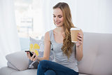 Young woman sitting on couch holding disposable cup texting on the phone