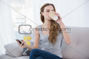 Young woman sitting on sofa holding disposable cup texting on the phone