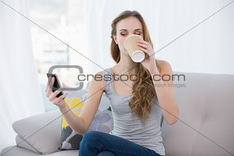 Young woman sitting on sofa drinking from disposable cup texting on the phone