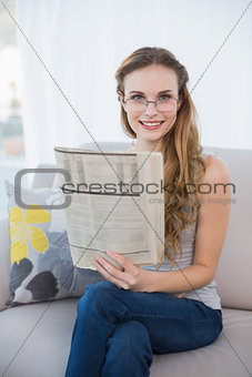 Cheerful young woman sitting on sofa holding newspaper