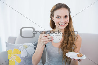 Cheerful young woman sitting on sofa holding cup and saucer