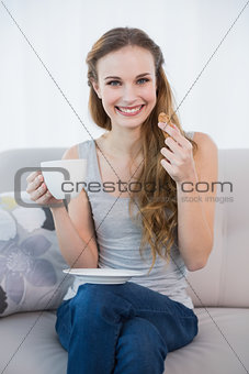 Cheerful young woman sitting on sofa holding cup and cookie