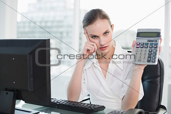 Frowning businesswoman showing calculator sitting at desk