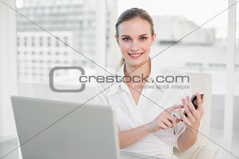 Smiling businesswoman using laptop and texting on smartphone