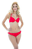 Half smiling blonde model posing with hands on hips in red bikini