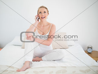 Natural cheerful blonde phoning while sitting on bed
