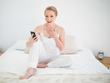 Natural surprised blonde holding smartphone while sitting on bed