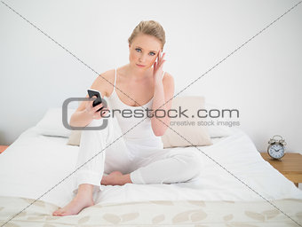 Natural serious blonde holding smartphone while sitting on bed