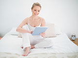 Natural smiling blonde using tablet while sitting on bed