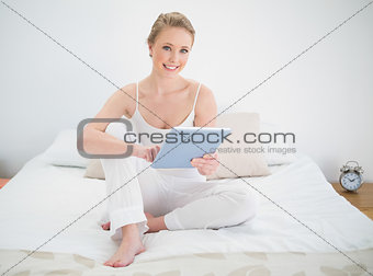 Natural smiling blonde holding tablet while sitting on bed