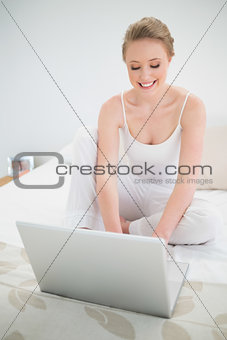 Natural cheerful blonde using laptop while sitting on bed