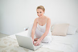 Natural smiling blonde using laptop while sitting on bed