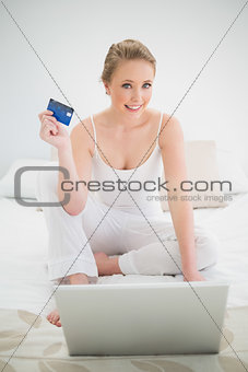 Natural smiling blonde holding credit card and sitting in front of laptop