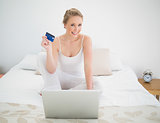 Natural cheerful blonde holding credit card and sitting in front of laptop