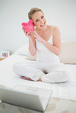 Natural cheerful blonde holding piggy bank while sitting on bed