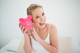Natural excited blonde holding piggy bank while sitting on bed